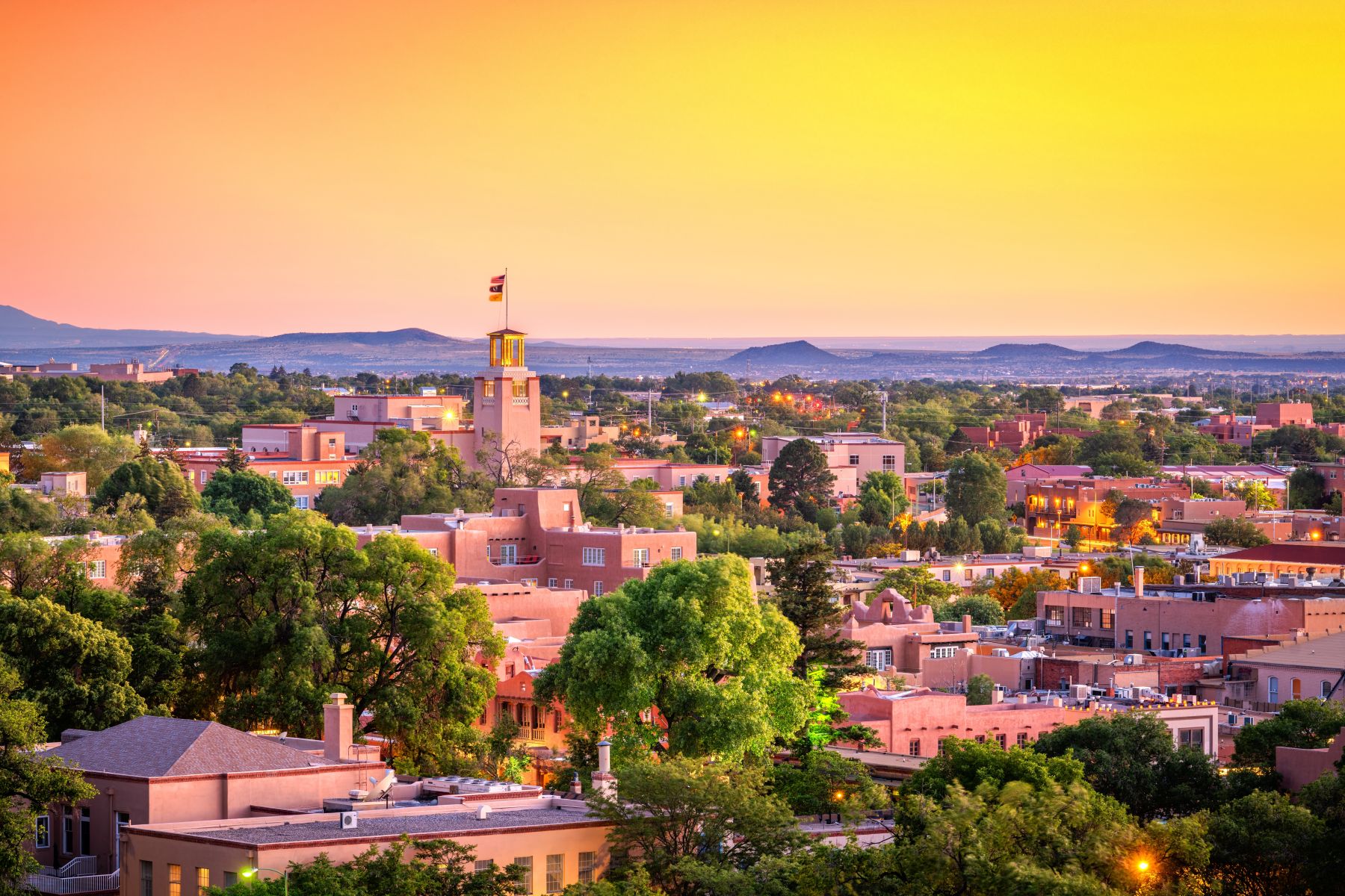 A breathtaking aerial view of the Santa Fe skyline during a golden sunset, featuring the iconic Pueblo-style architecture and the New Mexico State Capitol "Roundhouse" area, a historic backdrop for guests of Casas de Santa Fe vacation rentals.