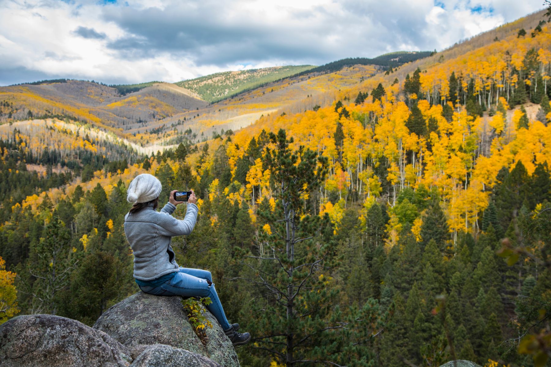 Person in a gray jacket and white knit hat sits on rocks, taking a photo of a forested mountainside with bright yellow autumn trees.